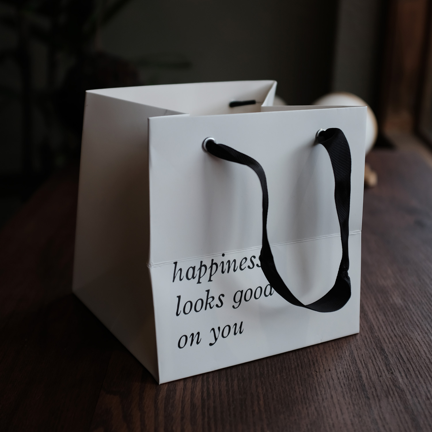 White paper bag with black handles on a wooden table, displaying the text 'happiness looks good on you'.