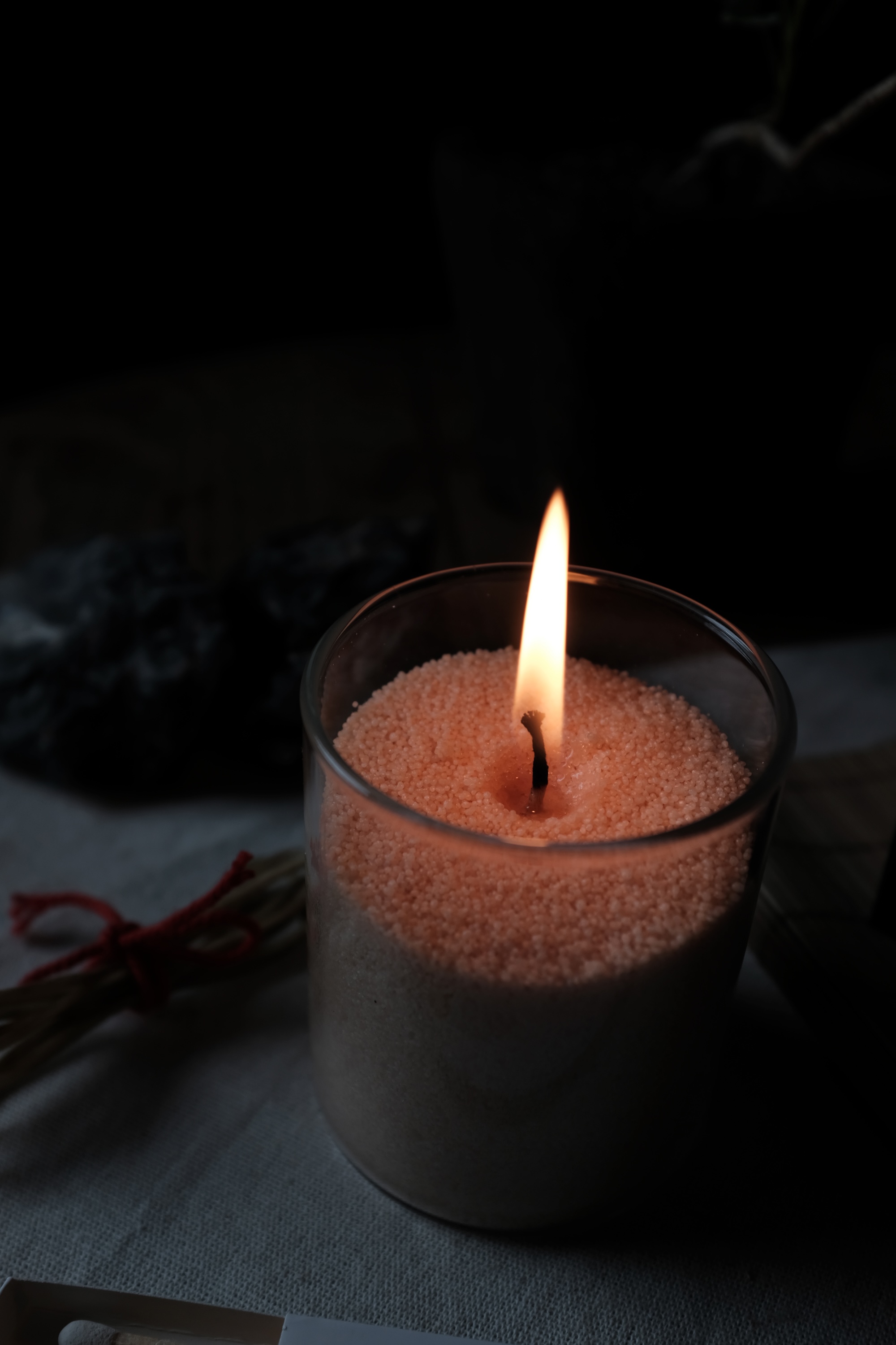 Lit candle in a glass container filled with small granules, placed on a cloth surface in a dimly lit setting.