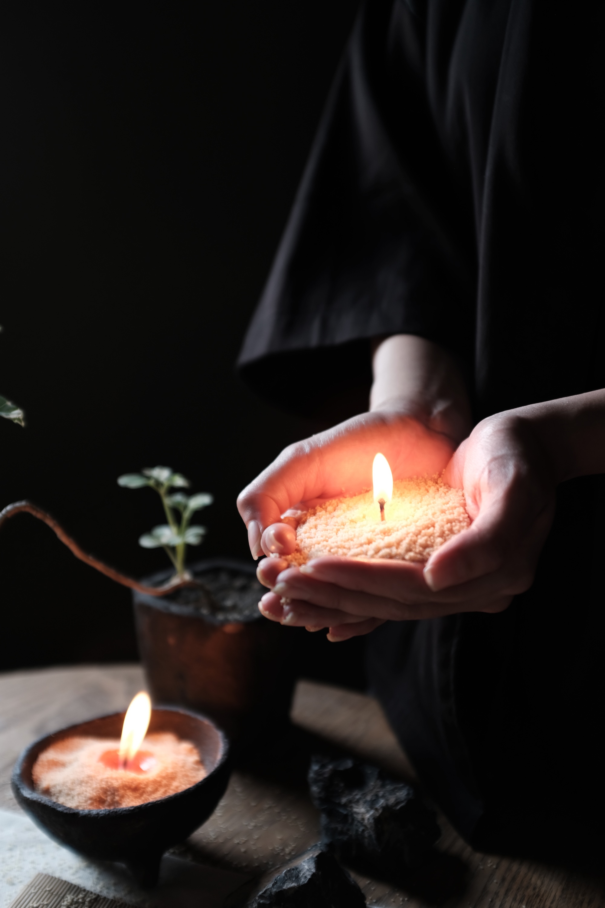 Hands holding a lit candle made of textured wax with a small green plant and another lit candle on a wooden surface in the background.