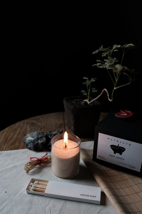 Lit sand candle in a glass container next to a potted plant, black box labeled 'sand candle', and a white matchbox labeled 'GOOD DAY' on a wooden table.