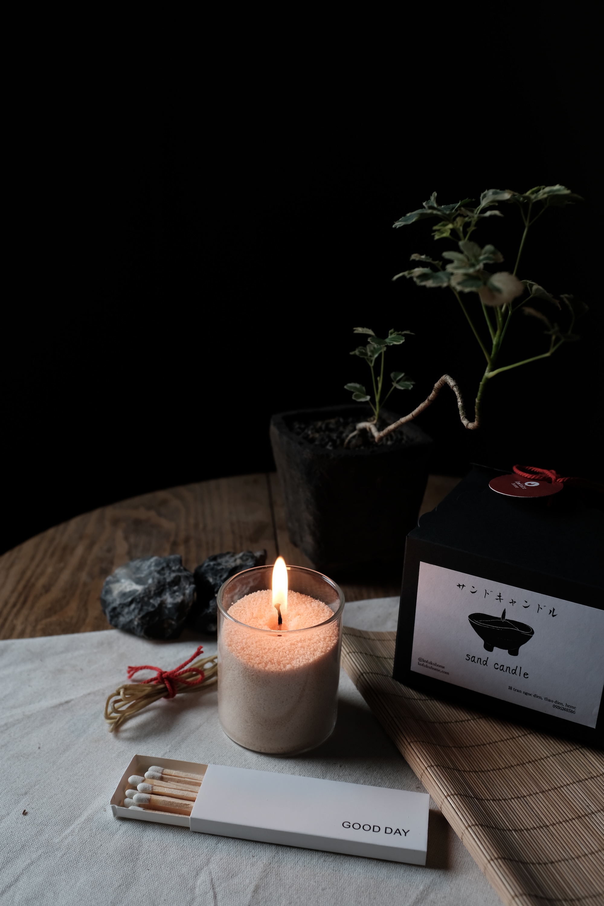 Lit sand candle in a glass container next to a potted plant, black box labeled 'sand candle', and a white matchbox labeled 'GOOD DAY' on a wooden table.