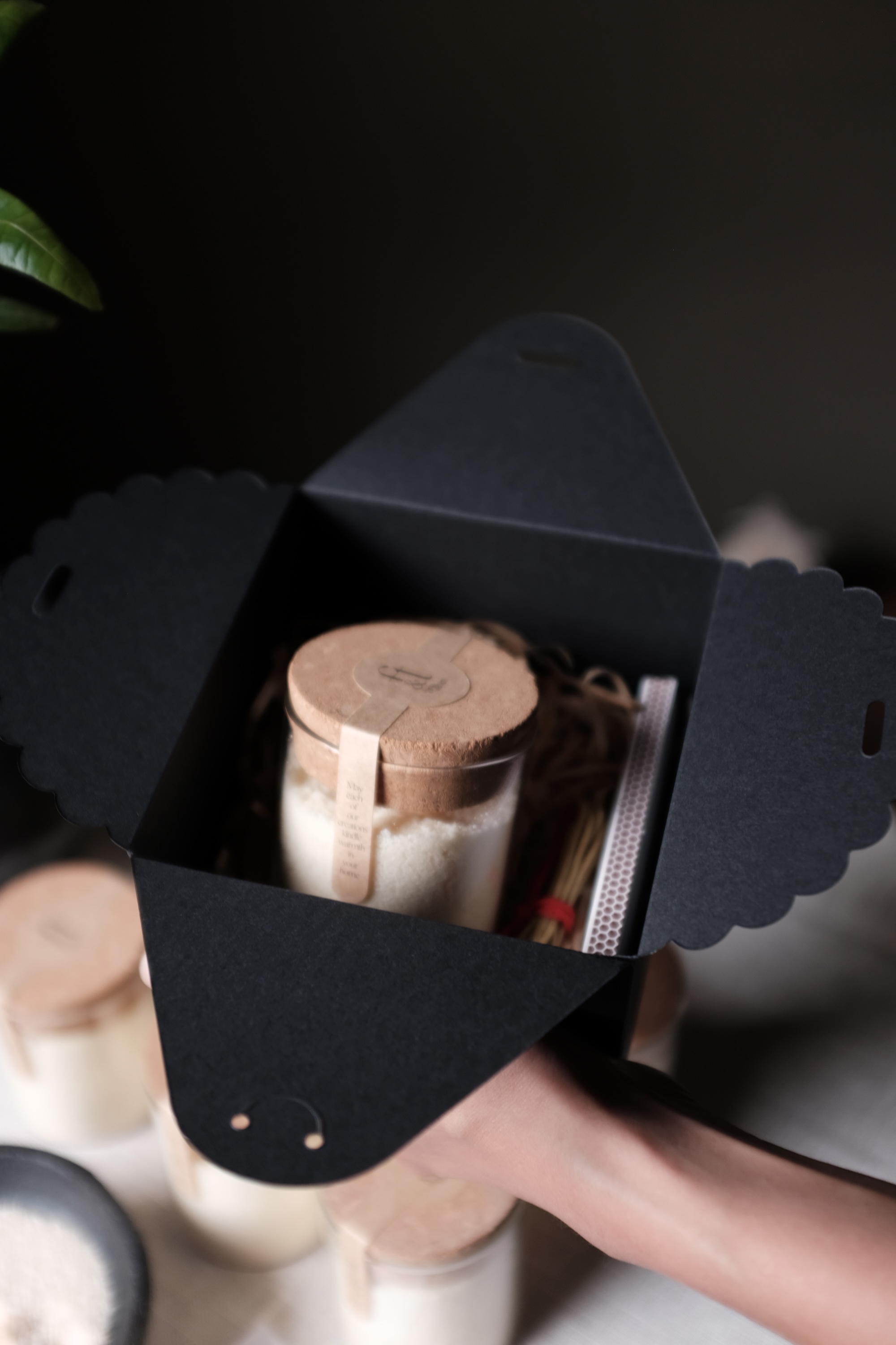 Hand holding a black gift box containing a glass jar with a cork lid and a small bundle tied with a red band.