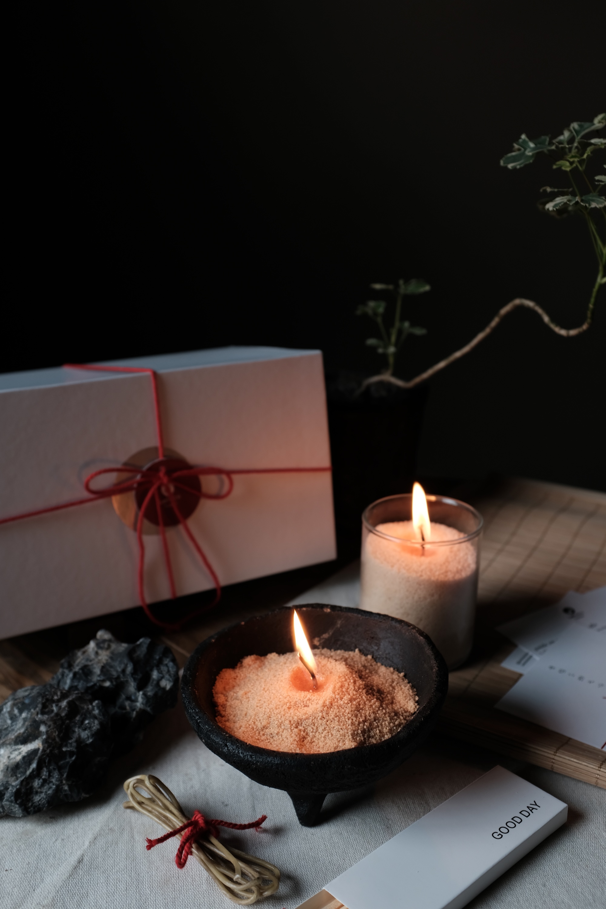 Two small burning candles surrounded by a white gift box tied with red string, a black rock, a plant, and a bundle of thin twine on a table.