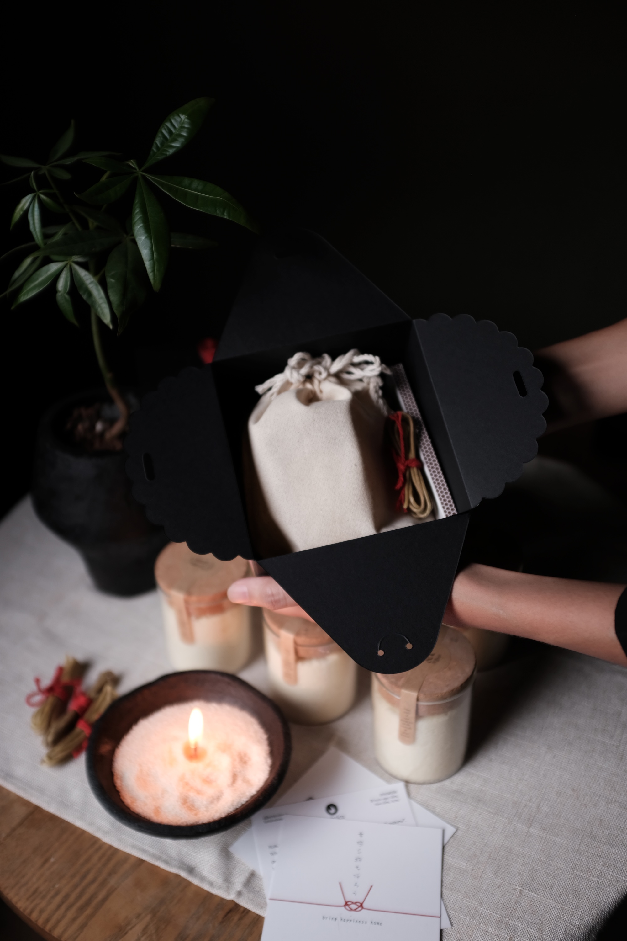 Hands holding an open black gift box containing a white drawstring pouch, with a lit candle, three jars, a small plant, and envelopes on a table.