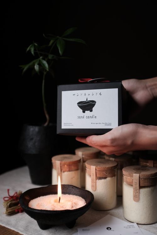 A lit sand candle in a black bowl on a table with several corked jars of candle sand and hands holding a black box labeled 'sand candle' in the background.