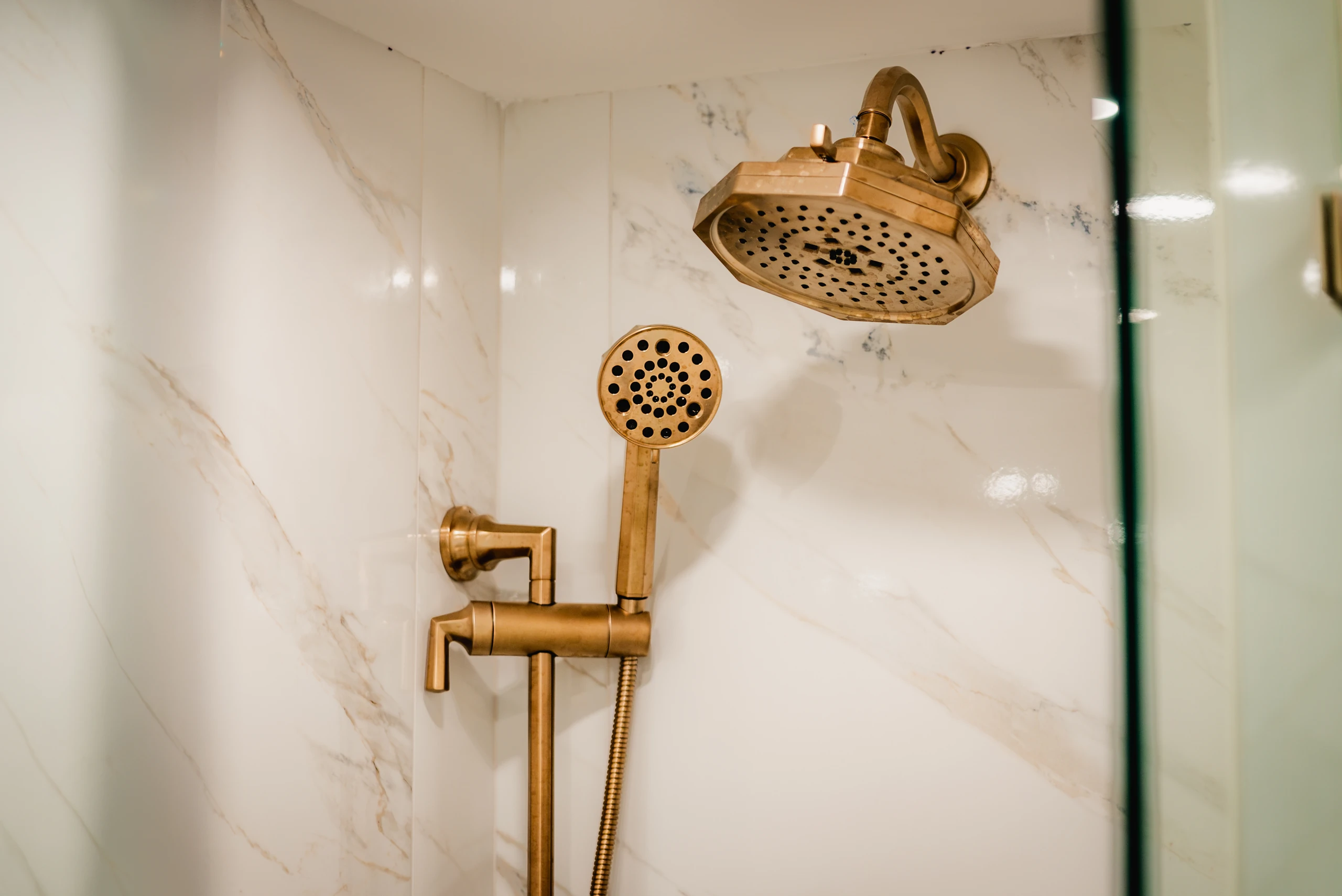 Brass rain showerhead and handheld sprayer on a marble-look shower wall