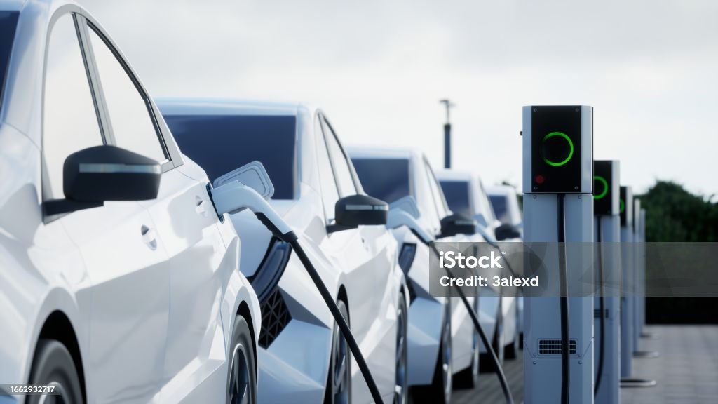 Row of white electric cars plugged into charging stations with green lights outdoors.