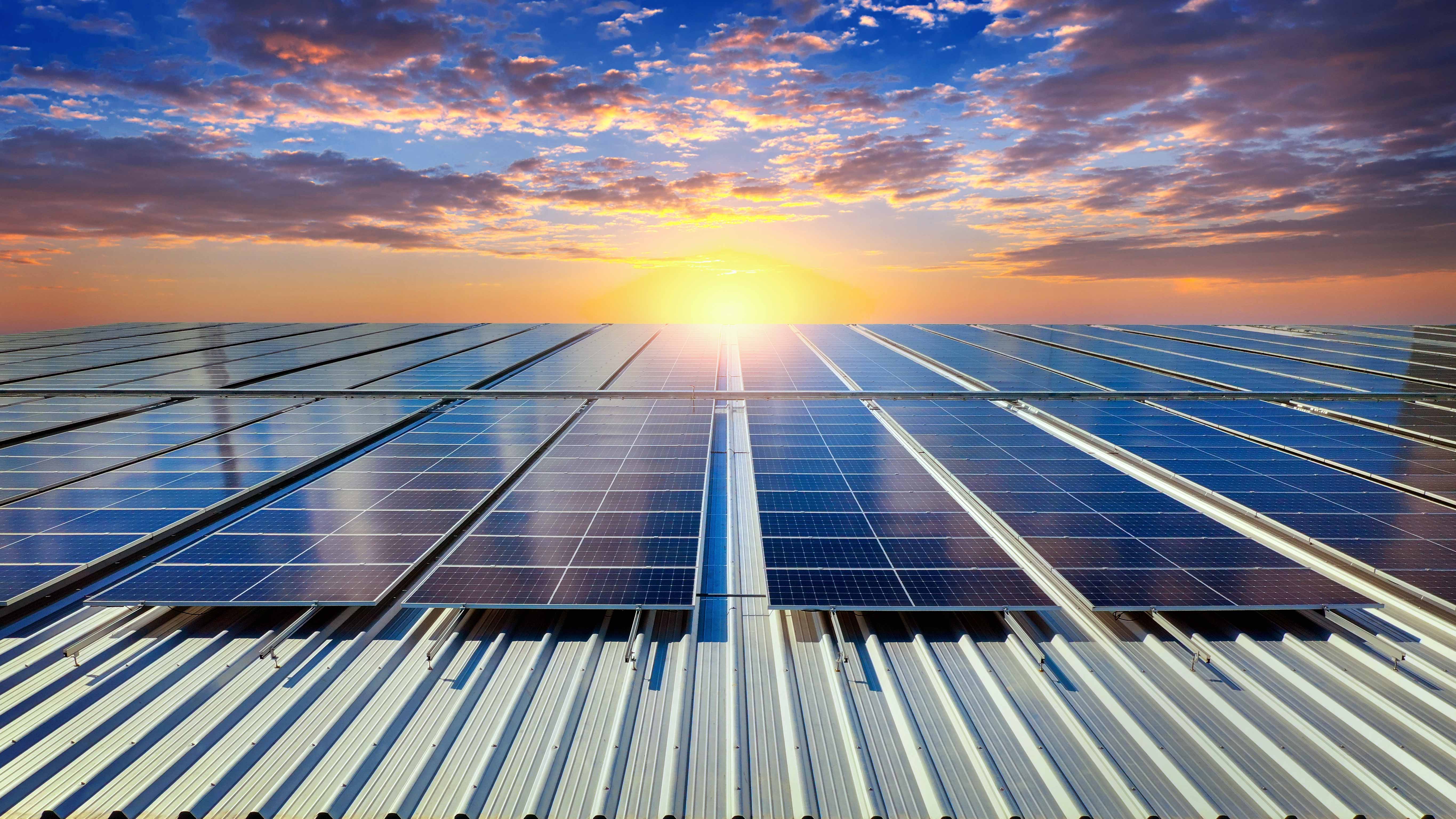 Rows of solar panels on a metal roof with a vibrant sunrise and partly cloudy sky in the background.