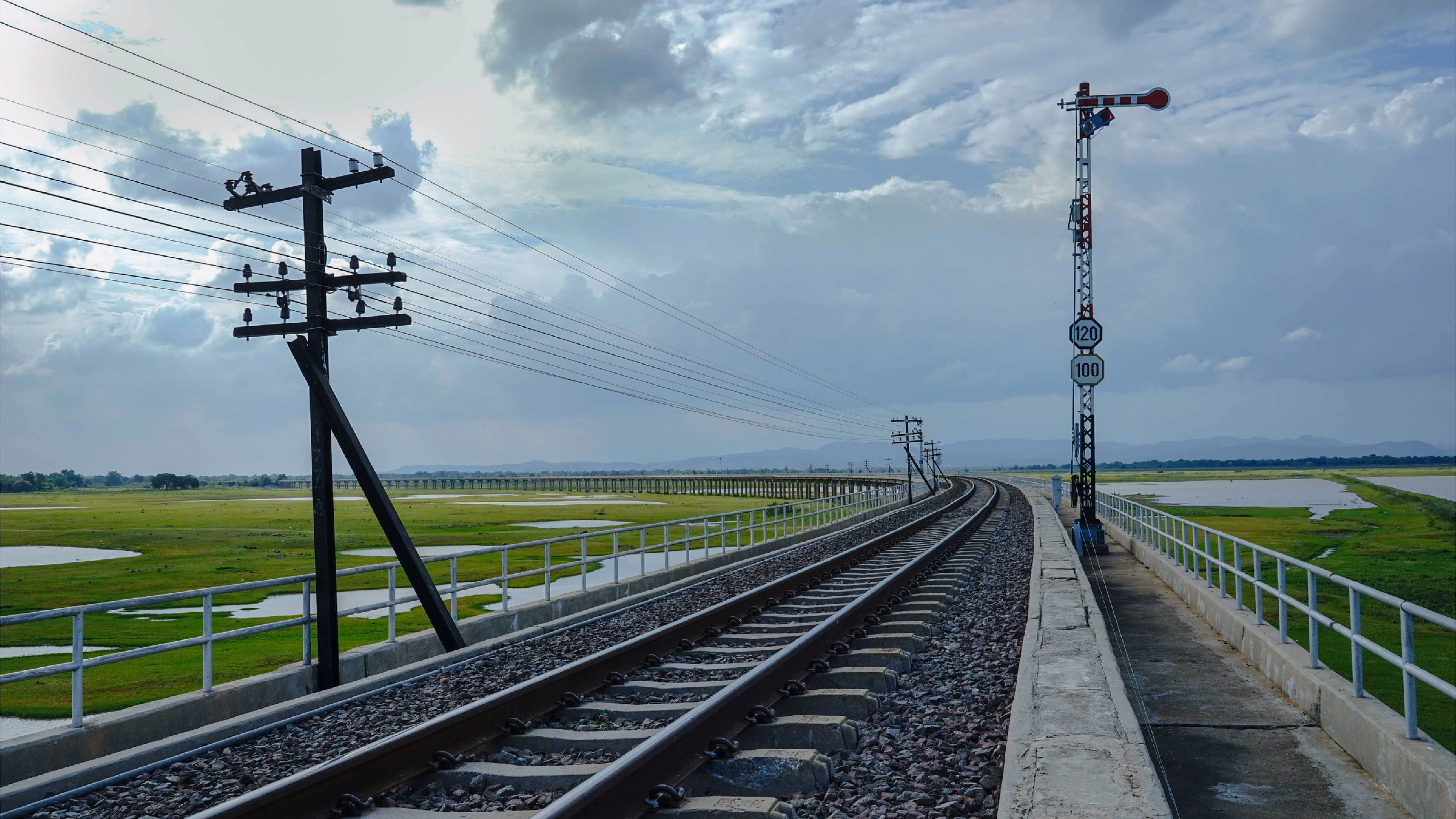 Railway tracks curving across a bridge with electric poles and a signal under cloudy sky.