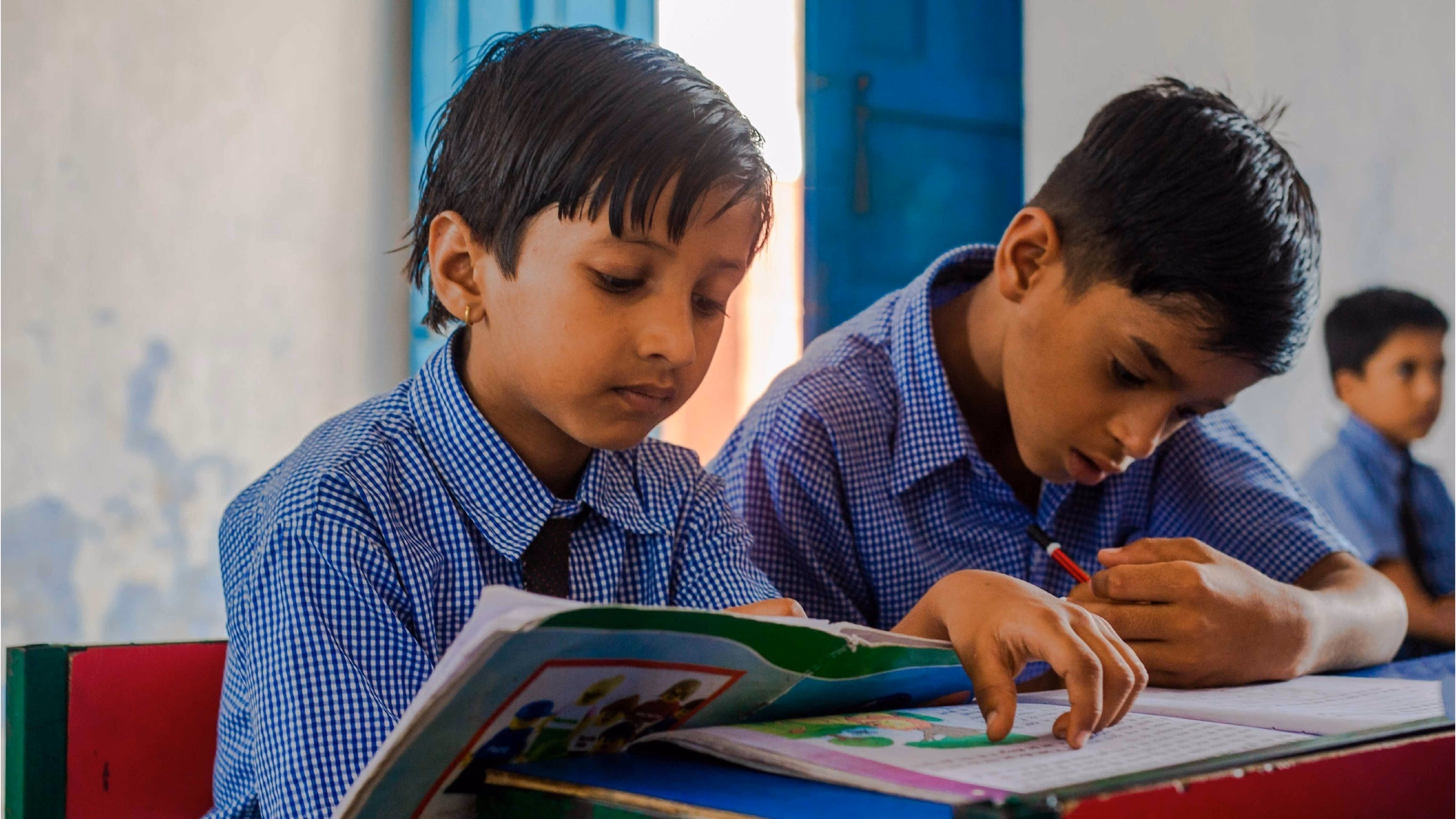 Two boys in blue checkered shirts studying and writing in notebooks at a classroom desk.