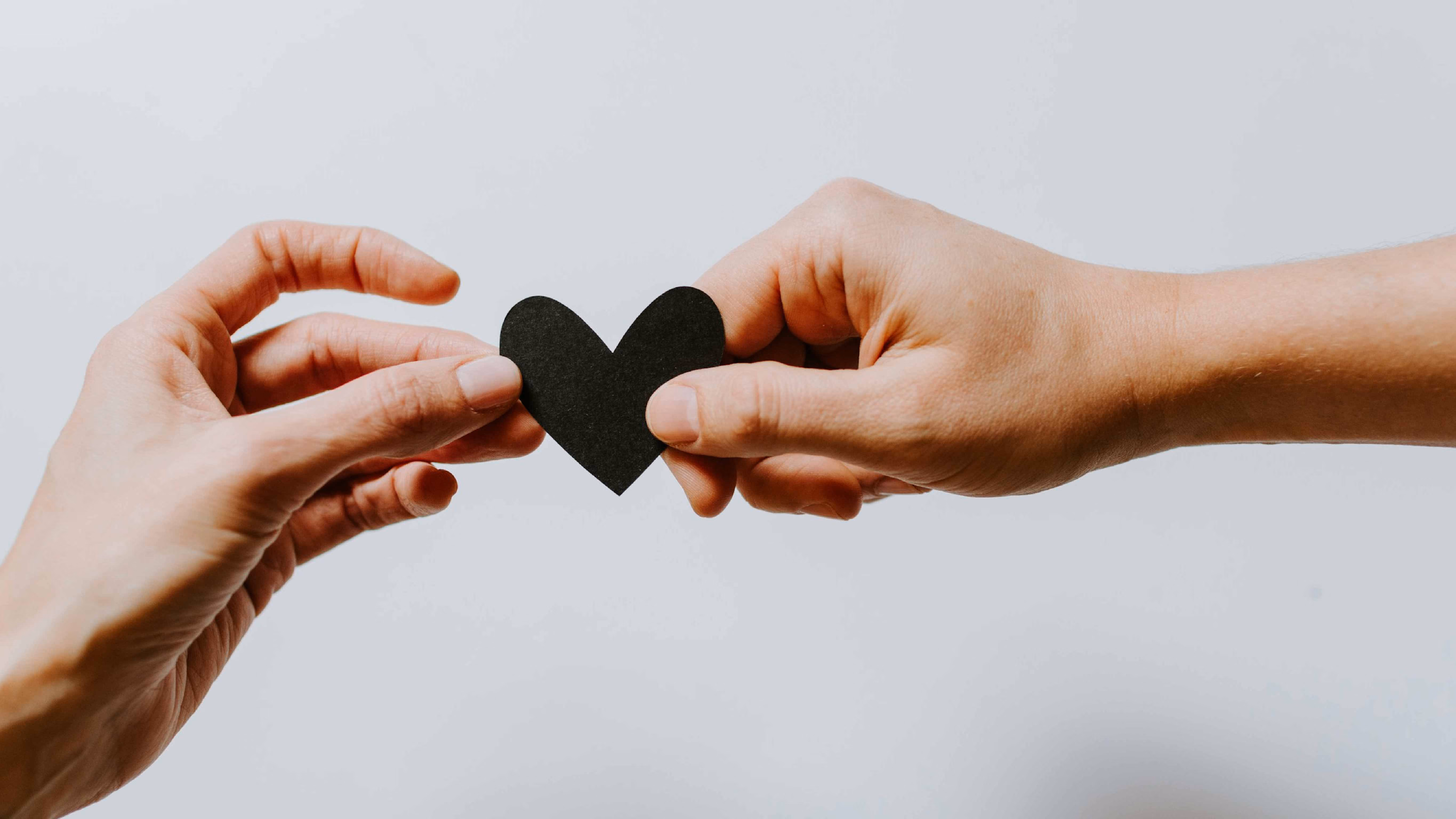 Two hands holding opposite sides of a small black paper heart on a plain background.