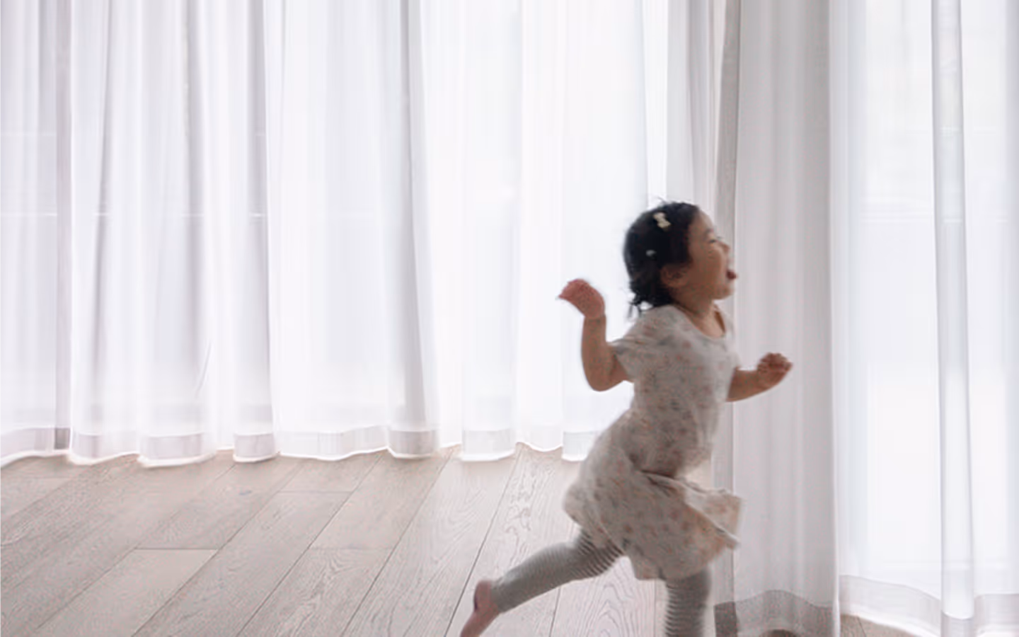 Young child in dress and leggings running happily beside sheer white curtains in a bright room.