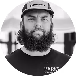 Man with beard wearing a Carhartt cap and a Parker Installation shirt in a blurred background.