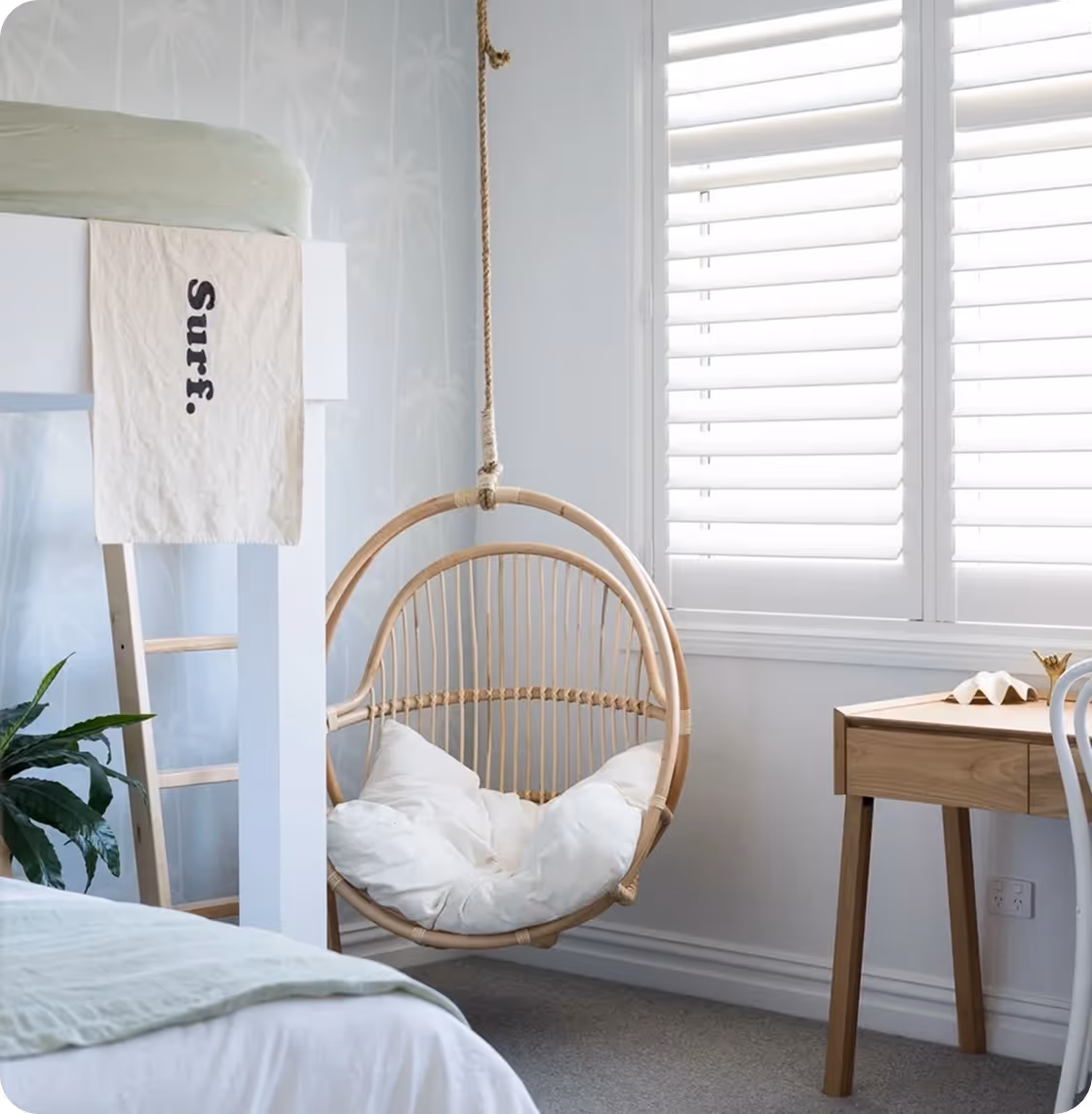 Light, airy bedroom with a hanging wicker chair, loft bed with Surf towel, wooden desk, and window blinds.
