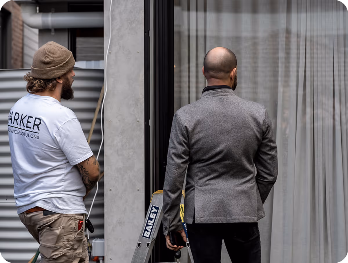 Two men stand facing a building window; one in work clothes and the other in a gray jacket.