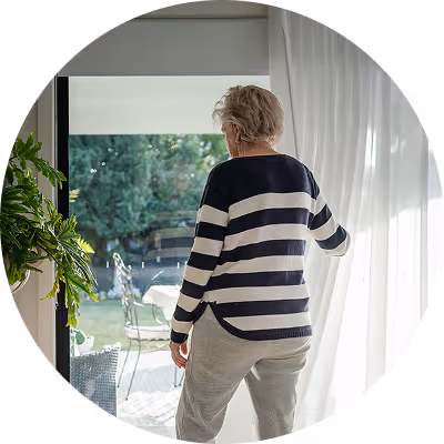 Older woman wearing striped sweater opening white curtains in bright room overlooking outdoor patio.