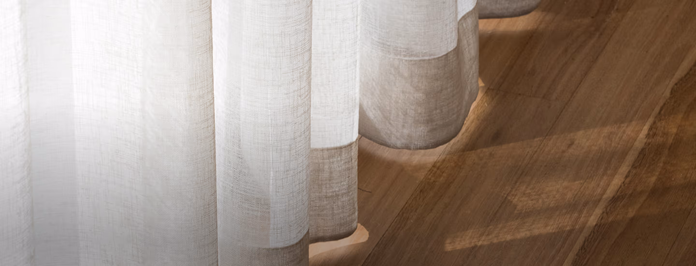 Light beige sheer curtains hanging above wooden floor with sunlight casting soft shadows.