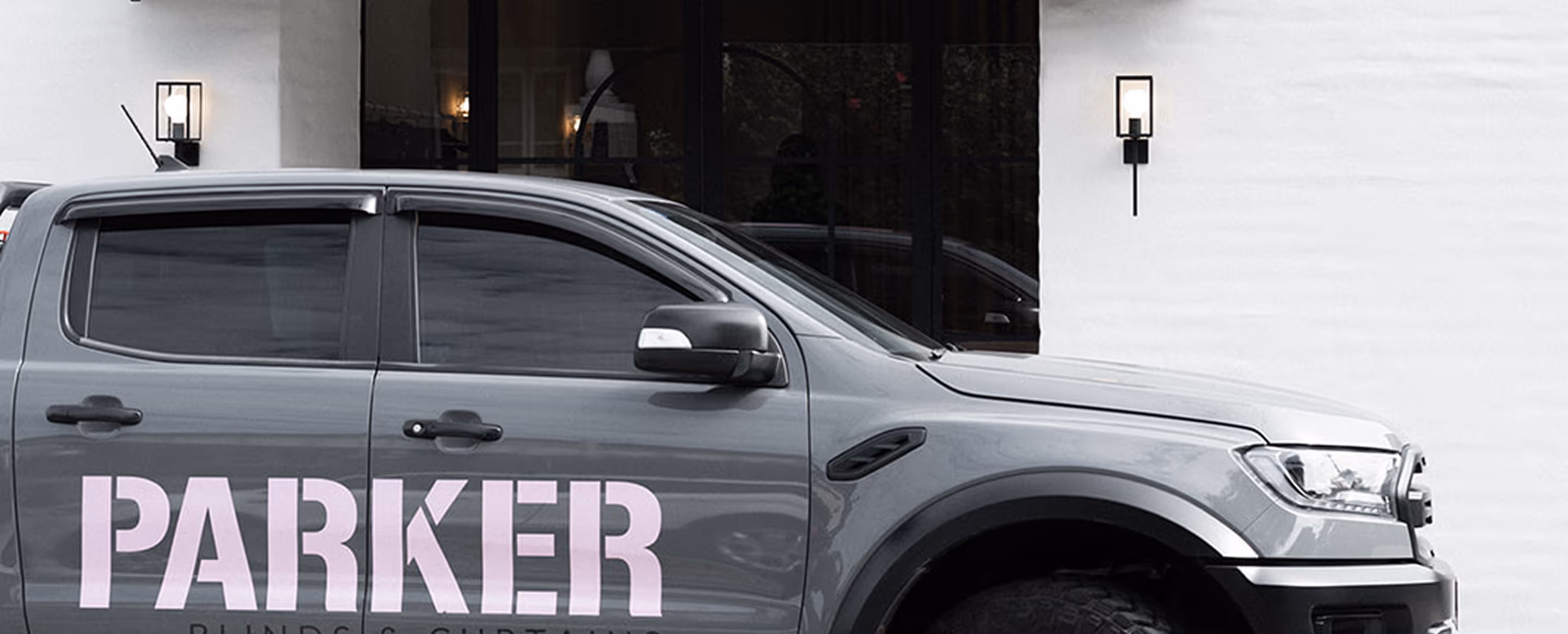 Gray pickup truck with the word PARKER in bold light pink letters on the side parked by a white building.