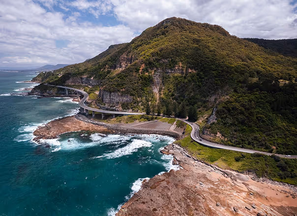 Curvy coastal road hugs rocky shore with ocean waves and a large forested hill.