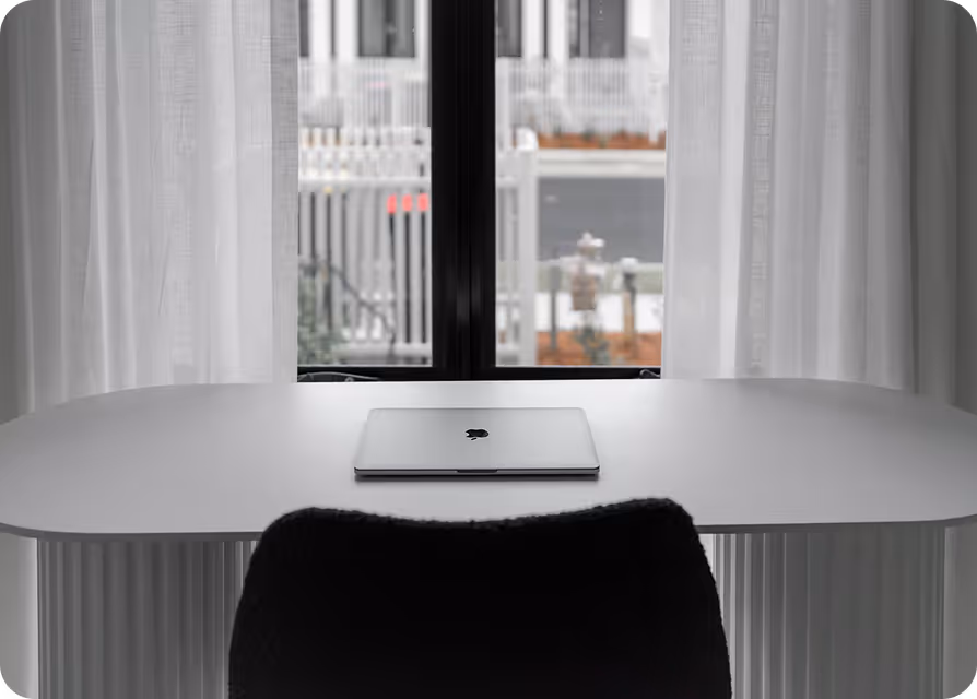 Closed laptop on a white desk with a black chair in front of a window with sheer white curtains.
