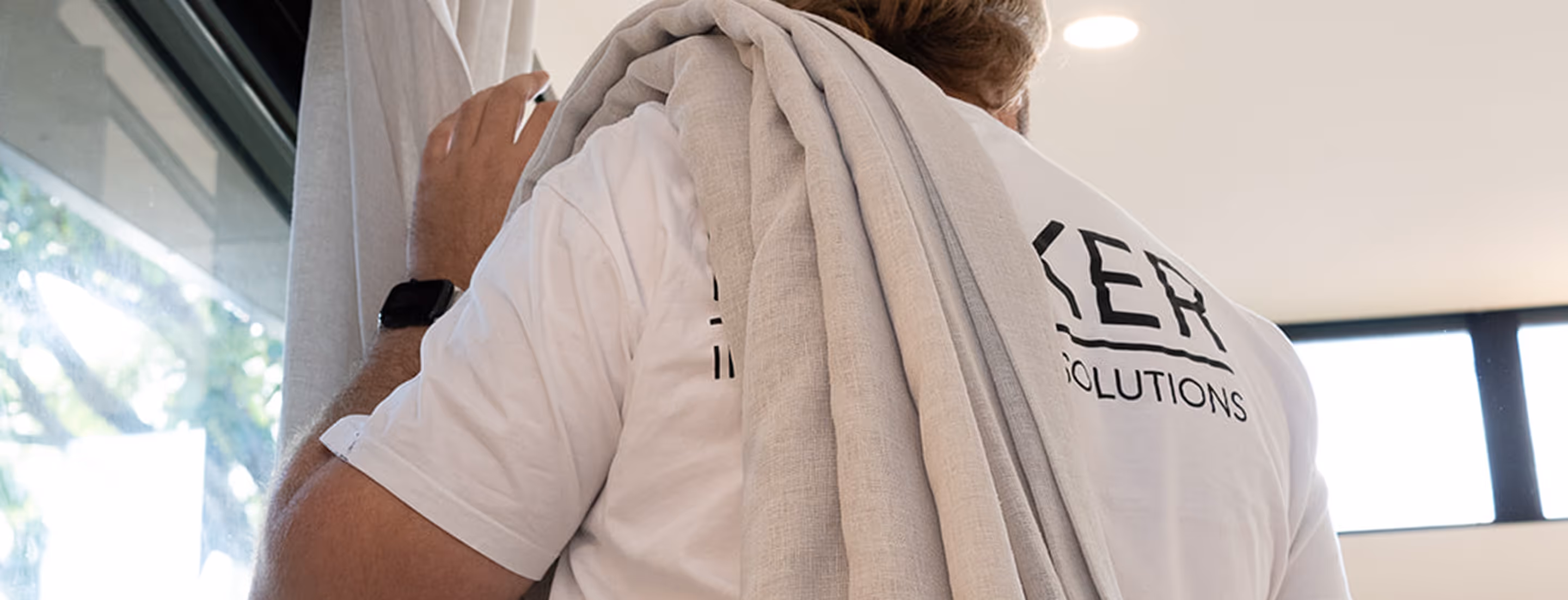 Man wearing white shirt carrying rolled-up beige carpet over his shoulder indoors near a window.