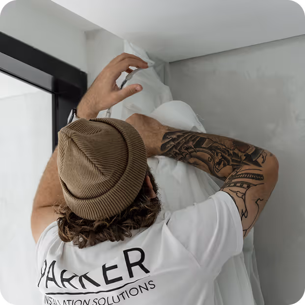 Man wearing a brown beanie installs white insulation material on a gray wall near a window.