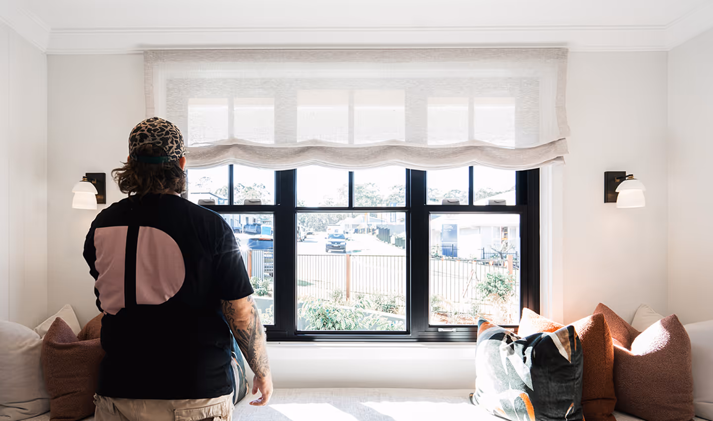 Person standing in front of large window with sheer blinds and cushions on a bench below.