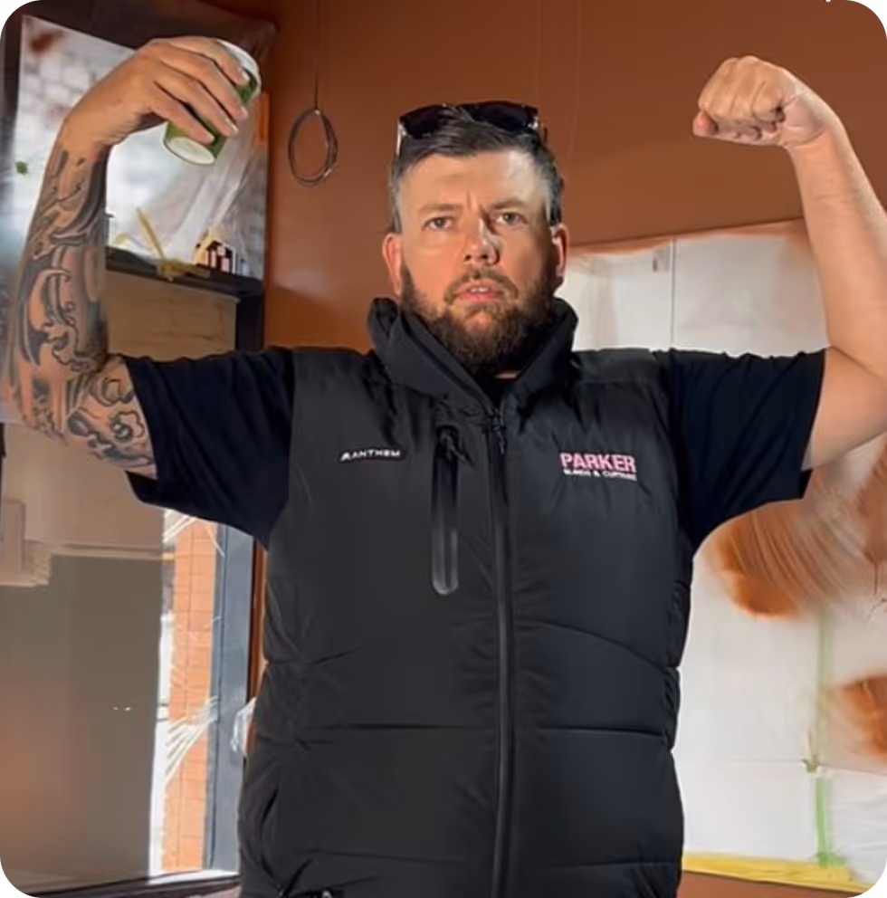 Bearded man flexes muscles, wearing sunglasses on head and a black vest with Parker logo.
