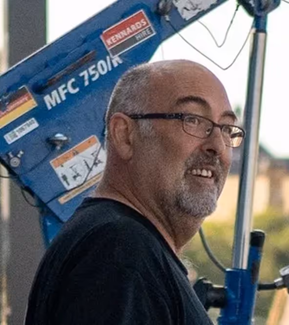 Smiling bald man with glasses and gray beard wearing a black shirt near blue machinery labeled MFC 750/K.