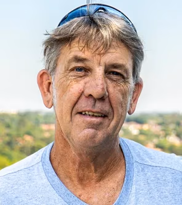 Older man with gray hair and sunglasses on his head smiling outdoors with a blurred green landscape behind him.