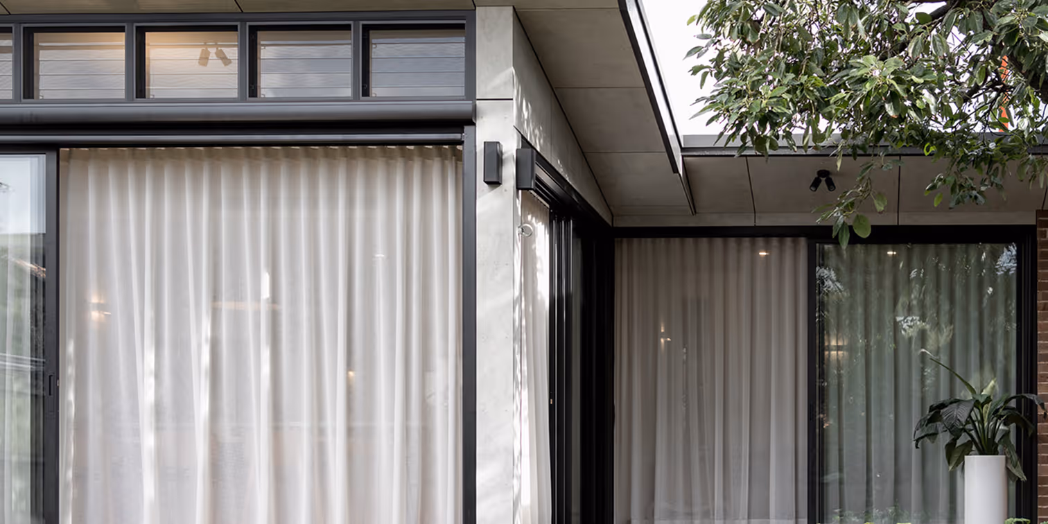 Modern house exterior with large black-framed windows covered by white curtains and a visible leafy tree branch.
