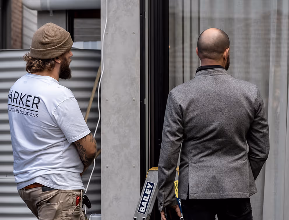 Two men stand facing a building; one in a white shirt and beanie, the other in a gray jacket.
