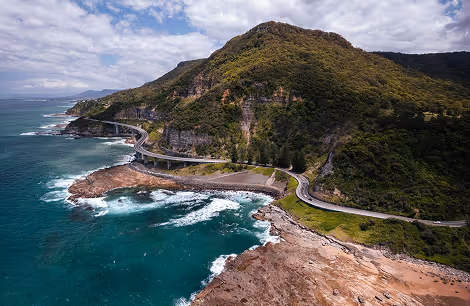 Coastal road winding around green hills beside rocky shore with waves crashing in ocean.