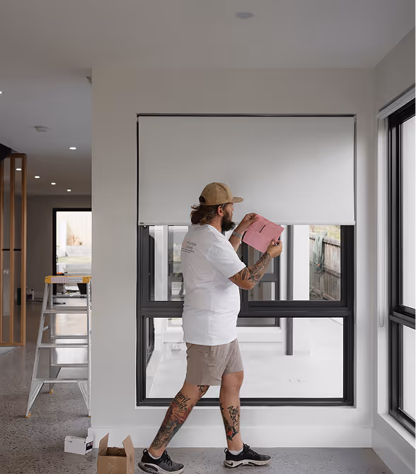 Man with tattoos adjusting a white window blind inside a modern house with large windows.