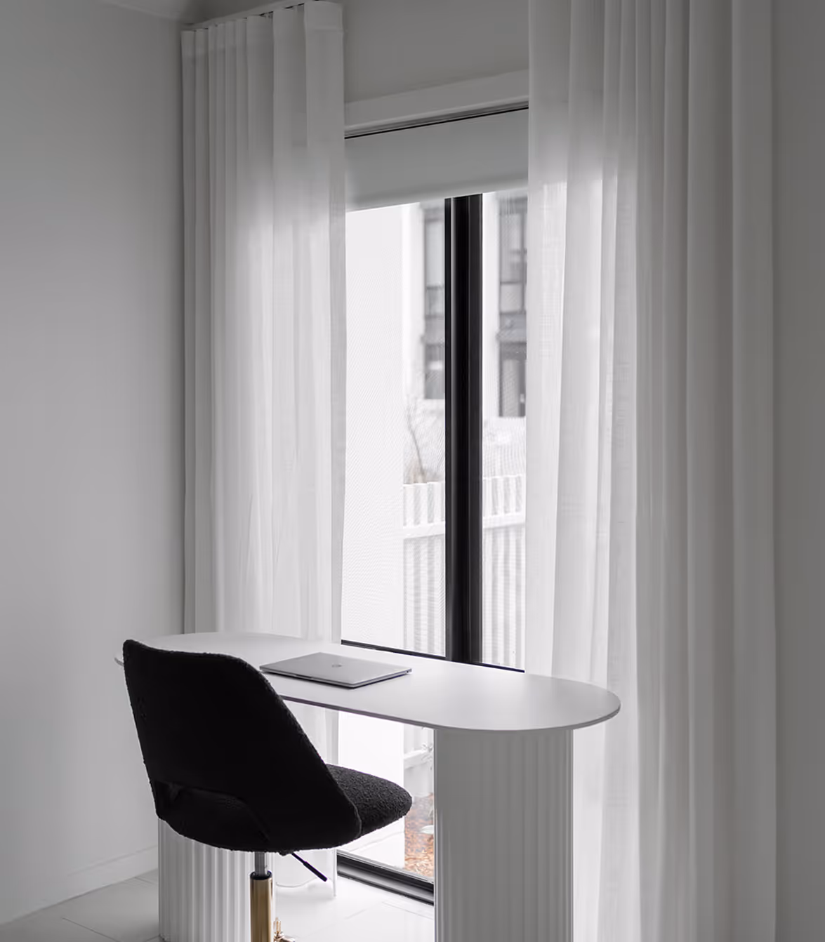 Minimalist white desk with closed laptop and black chair in front of a window with sheer white curtains.