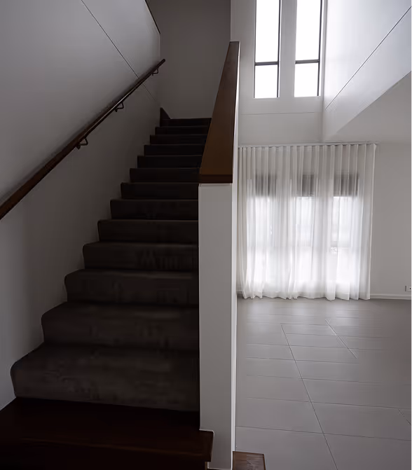 Modern indoor staircase with dark carpet on the left and sheer white curtains over large windows on the right.