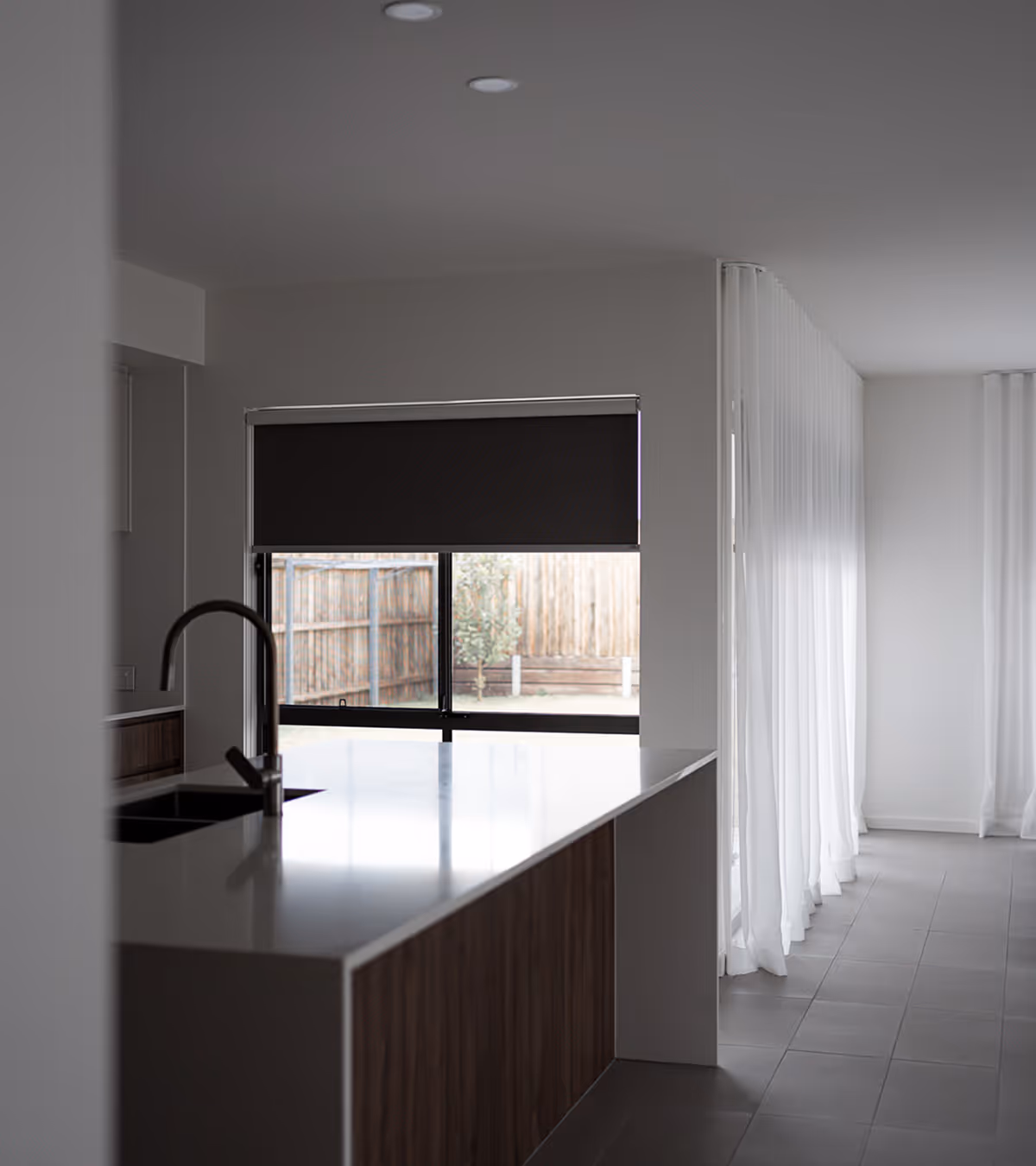 Modern kitchen with white countertop, dark wood cabinets, black faucet, and a window with dark roller blind.