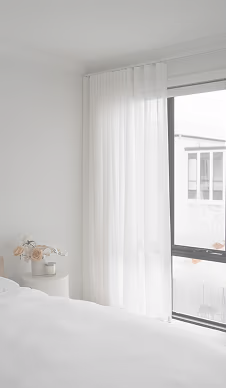 Bright bedroom corner with sheer white curtains, a small round table, and a vase of flowers.
