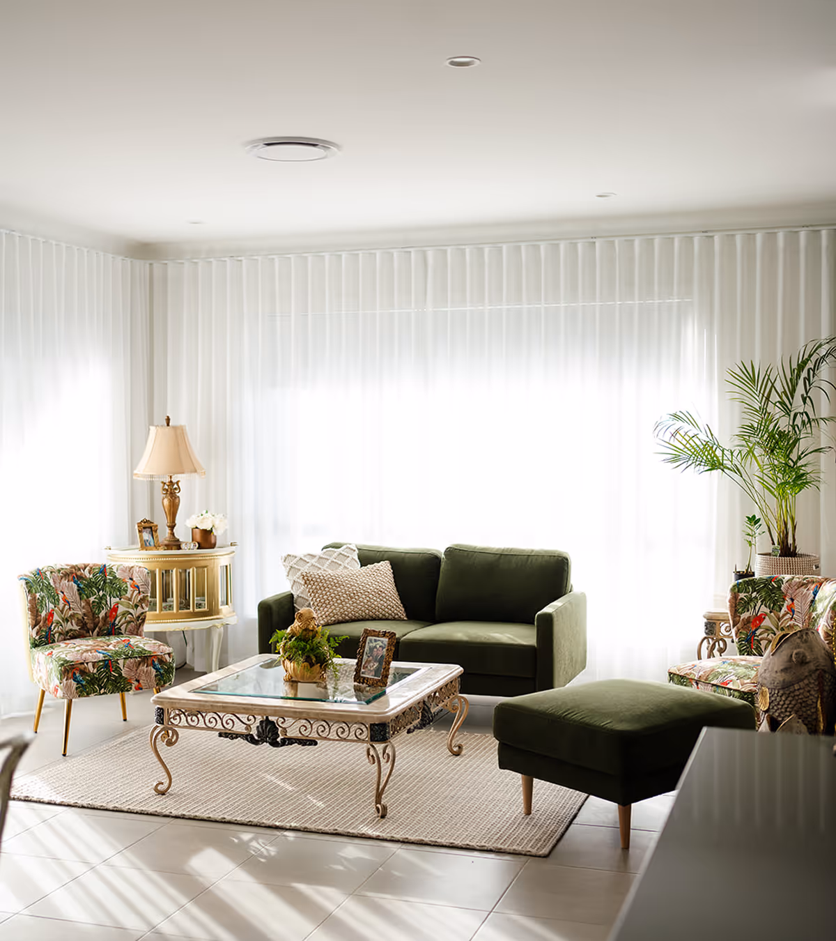 Bright living room with green sofa, matching ottoman, floral armchairs, glass-top coffee table, and plants.