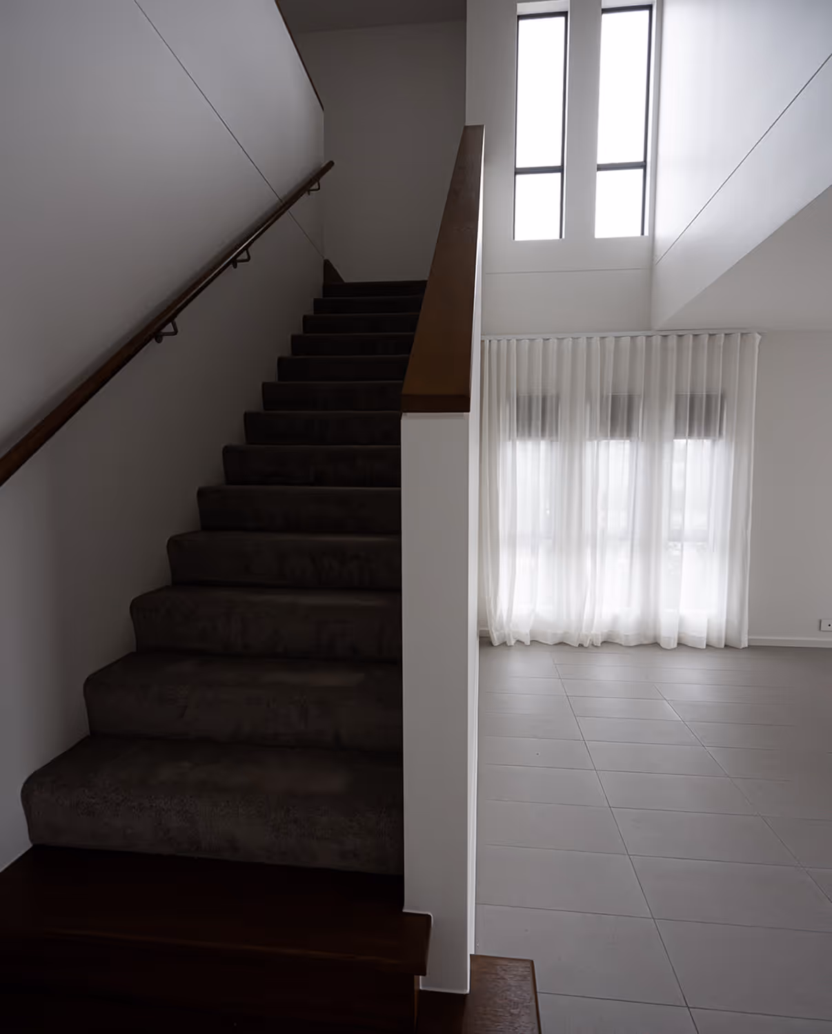 Interior view of carpeted stairs next to a bright window with sheer white curtains in a modern home.