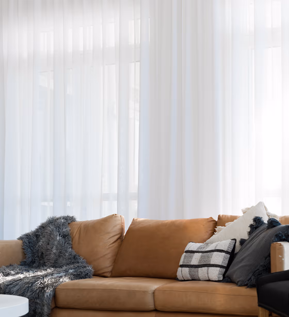 Brown leather sofa with gray and patterned pillows and a gray fluffy throw in front of sheer white curtains.