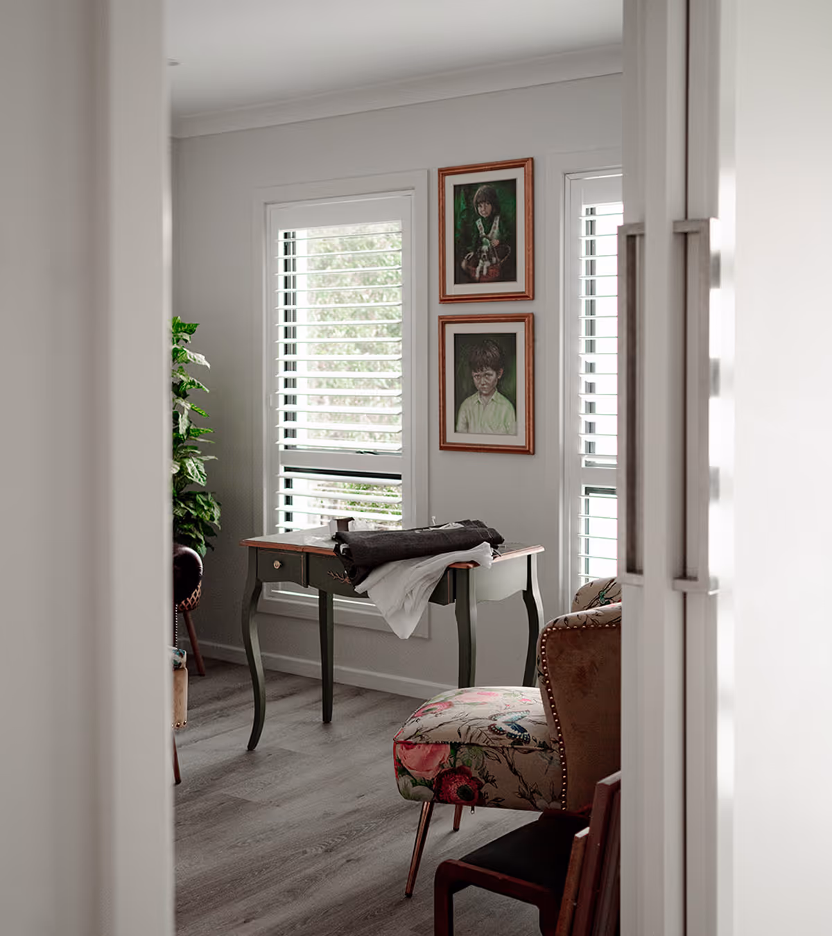 Room with wooden floor, shuttered windows, a dark green table holding clothes, and floral-upholstered chair.