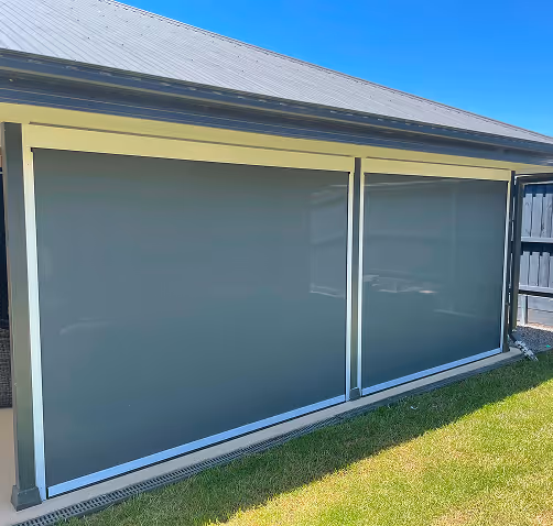 Outdoor roller shades covering two large windows on a house patio under a metal roof.