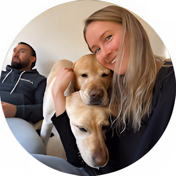 Smiling woman hugging two yellow Labrador retrievers while a man sits in the background.