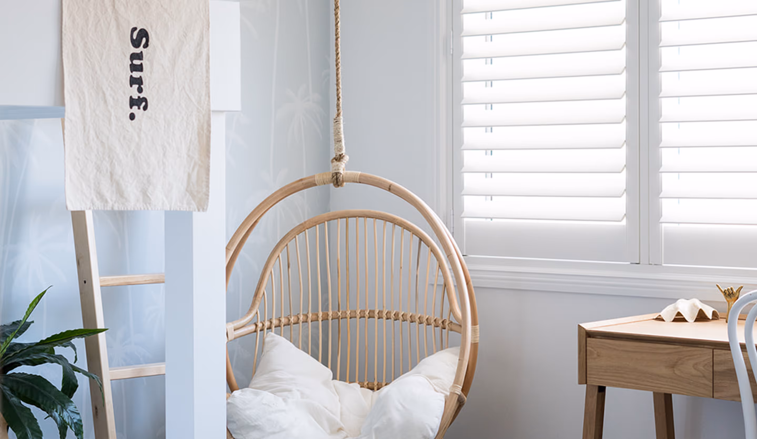 Light-filled room with hanging rattan chair, wooden desk, white shutters, and a ladder with a surf towel.