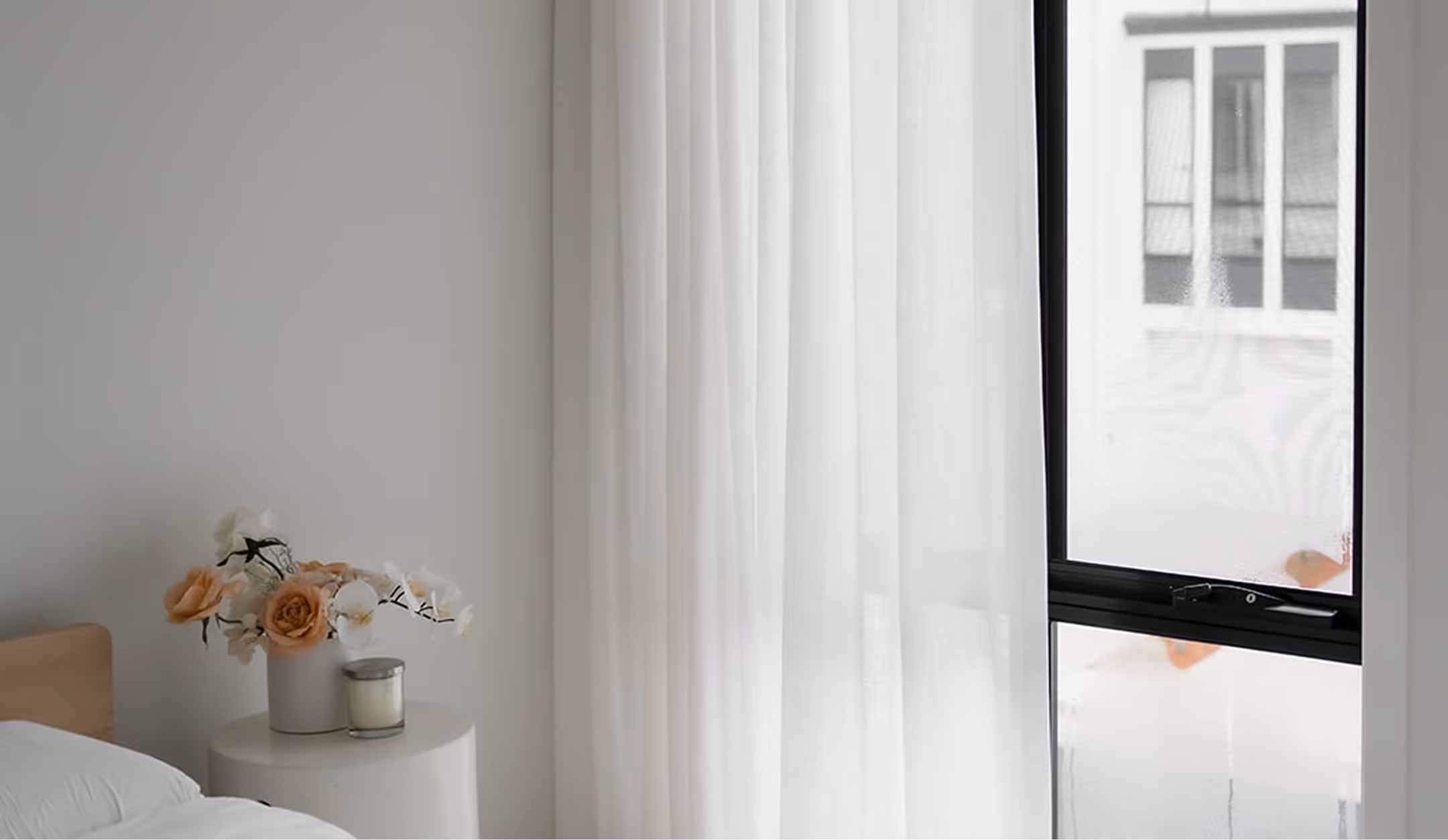 Minimalist bedroom corner with a white round table holding flowers and a candle beside sheer curtains and a window.