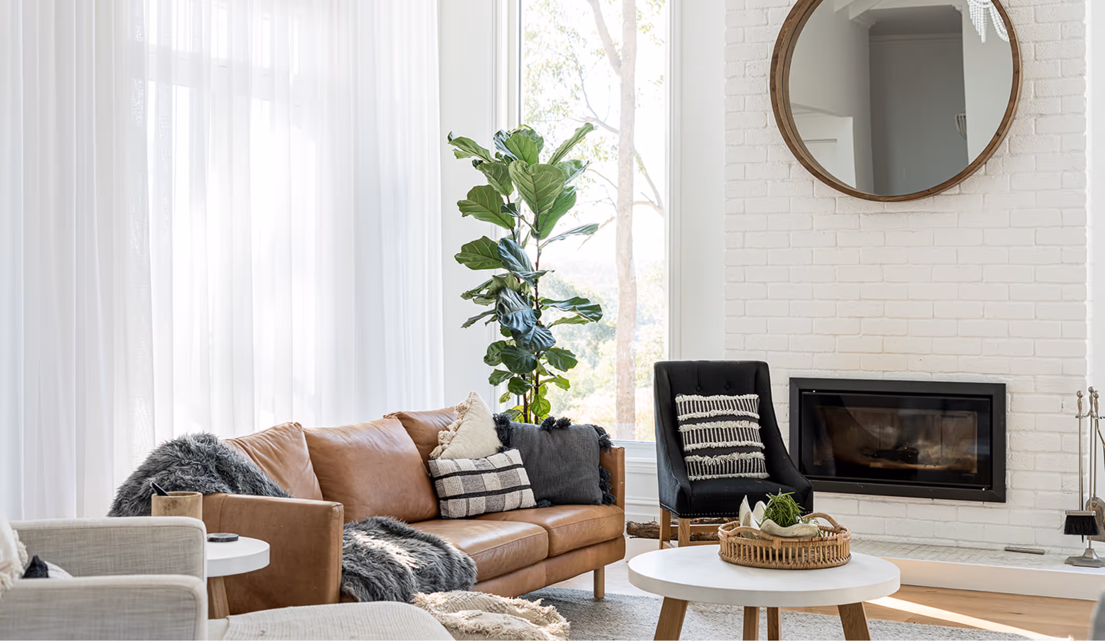 Modern living room with tan leather sofa, black chair, round white coffee table, and tall green plant.