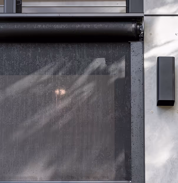 Black motorized roller curtain installed outside with water droplets on its surface.