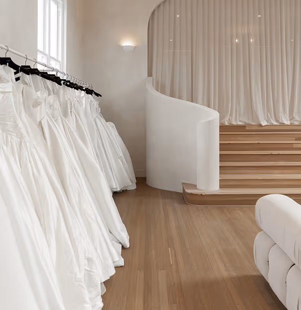 Row of white wedding dresses on black hangers in a minimal bridal boutique with wooden flooring and a curved staircase.