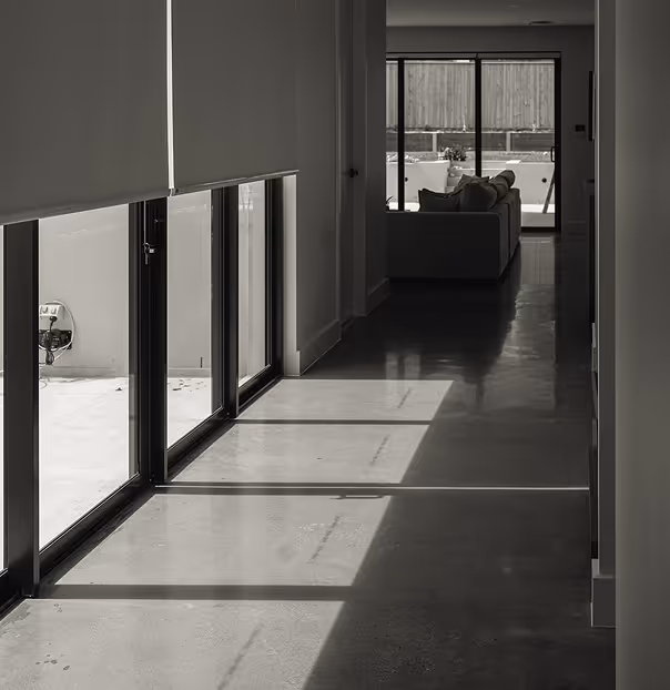 Sunlight casts window shadows on a polished floor in a minimalist hallway leading to a seating area.